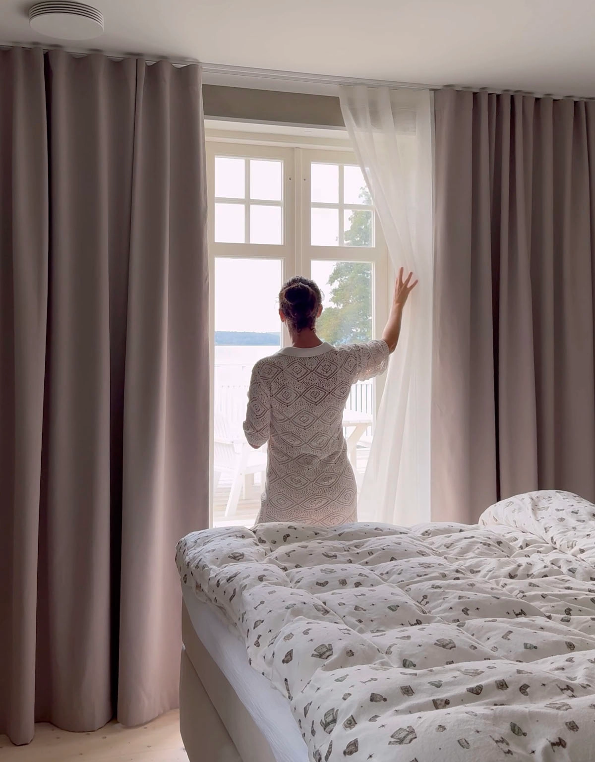 Kvinna som drar en vit skir gardin åt sidan, som hänger bakom beiga mörkläggningsgardiner