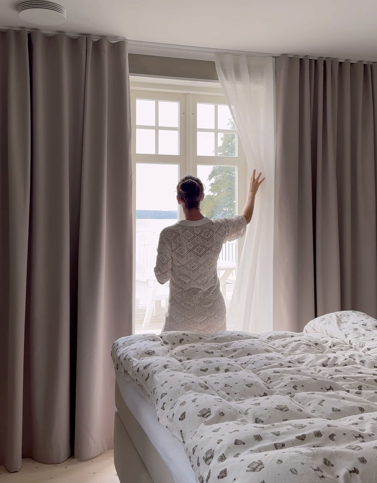 Kvinna som drar en vit skir gardin åt sidan, som hänger bakom beiga mörkläggningsgardiner